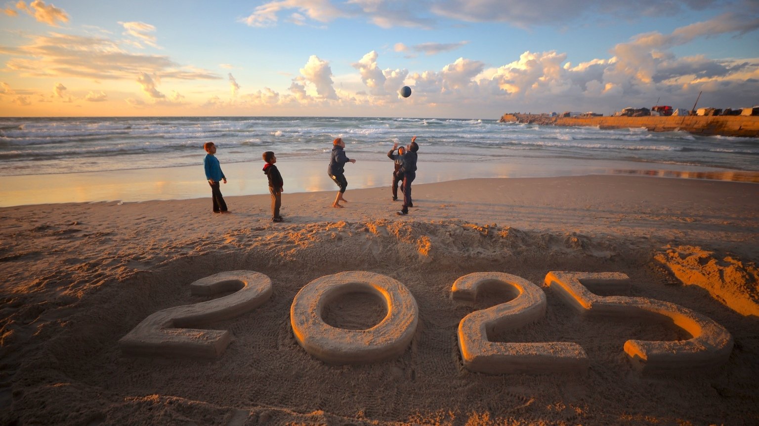 Children on a beach playing with a ball by a '2025' sand sculpture