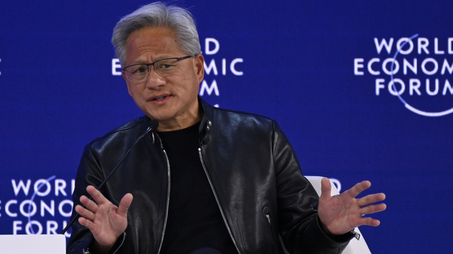 NVIDIA's Jensen Huang making a two handed wave gesture while talking on a World Economic Forum stage