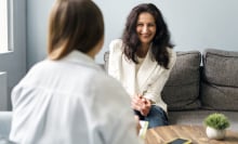 A female therapist talks to a woman sitting on a couch. A phone is on the table. 