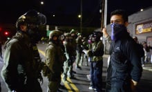 A line of masked and armed ICE officials stands facing a group of protesters.