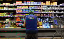 An employee in a blue vest kneels in front of a refrigerated section of a grocery store.