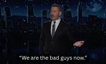 A man in a suit stands on a talk show stage. The caption at the bottom reads, "We are the bad guys now."