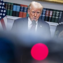 Trump stands at a podium surrounded by blurred out cameras and press members.