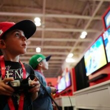 Two small children wearing Nintendo merch play a new Switch 2 game in front of large monitors. 