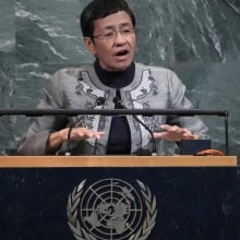 At the UN podium, a woman with short hair and glasses speaks. 