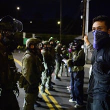 A line of masked and armed ICE officials stands facing a group of protesters.