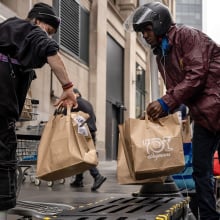 Two delivery drivers pass paper bags of groceries to each other on a city street.