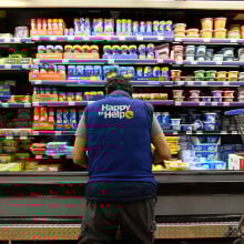 An employee in a blue vest kneels in front of a refrigerated section of a grocery store.