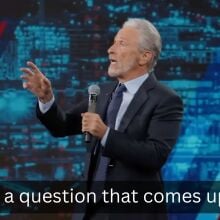 A man stands on a stage in a suit with a microphone, pointing out towards the audience.