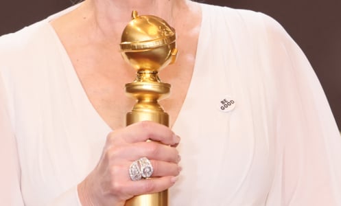 A close up image of Jean Smart holding a Golden Globe award. A white pin with the words "Be Good" is pinned to her chest.