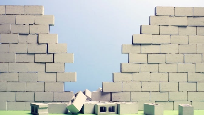 Concrete block wall with large breach in middle, several blocks laying on green surface