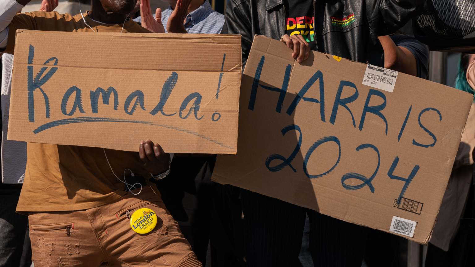 Two people each hold up a cardboard sign. One reads, "Kamala!" The other reads "Harris 2024".