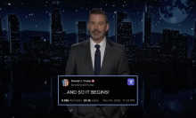 A man in a suit stands on a talk show stage. At the bottom of the screen is a Truth Social post from Donald Trump reading "...AND SO IT BEGINS."