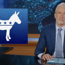 A man in a suit sits behind a talk show desk looking angry. In the top left is the logo of the Democrat party.