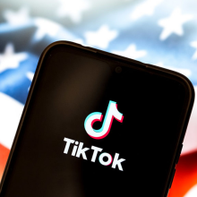 The TikTok logo appears on a smartphone screen, with the American flag on a computer screen in the background.