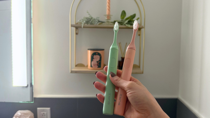 Hand holding pink and green Suri toothbrushes in front of bathroom wall